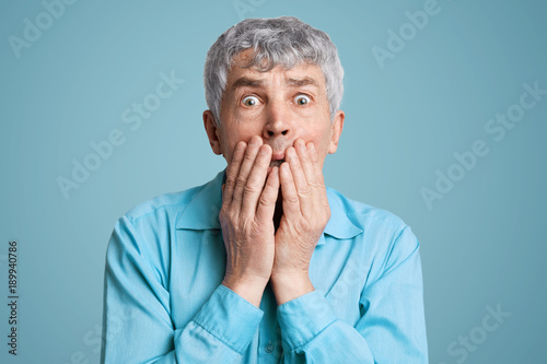 Stunned frightened elderly male model covers mouth with hands, being surprised and scared to hear shocking news, can`t believe in disaster, isolated over blue background. People and reaction