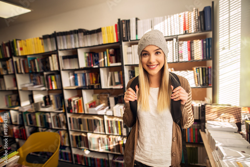 Portrait view of adorable happy attractive hardworking high school student girl standing in the sunny library or classroom and posing with backpack and hat while looking at the camera.