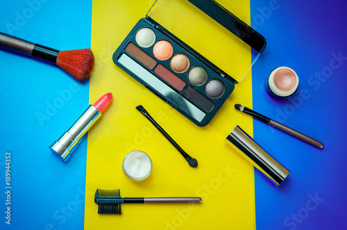 Cosmetic beauty products for make-up on bright colorful background – eye shadows, brushes, lipstick, face cream and highlighter base in small jars.