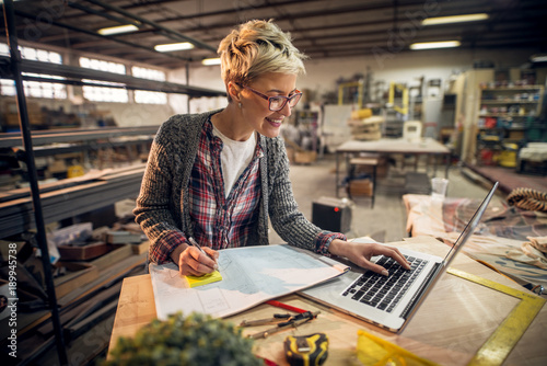 Close up view of charming smiling motivated short hair attractive middle aged industrial female engineer with eyeglasses working with blueprints and laptop in the workshop.
