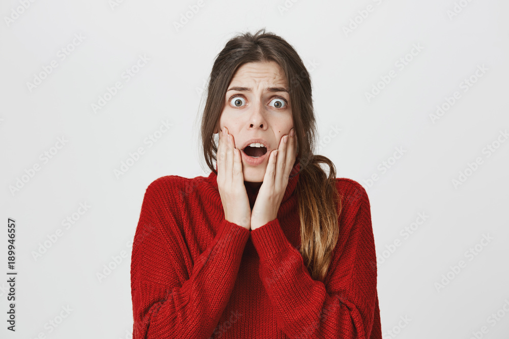 Frightened scared dark-haired woman with ponytail wears red sweater, looks with terrified expression, being afraid of something, poses against gray background with popped eyes and opened mouth