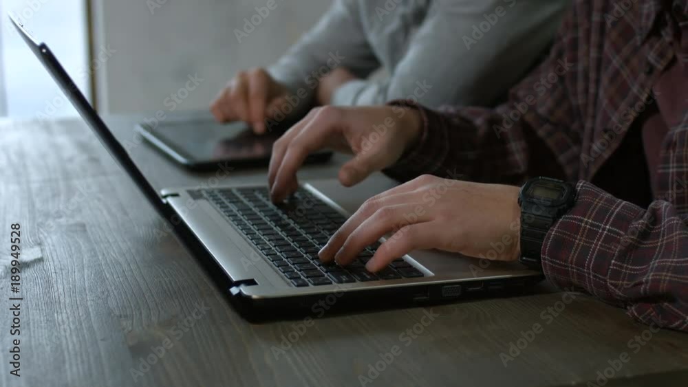 Closeup male hands typing on modern laptop keyboard and browsing the net on digital tablet pc on the wooden table. Side view. Young men working with laptop and tablet pc and networking.