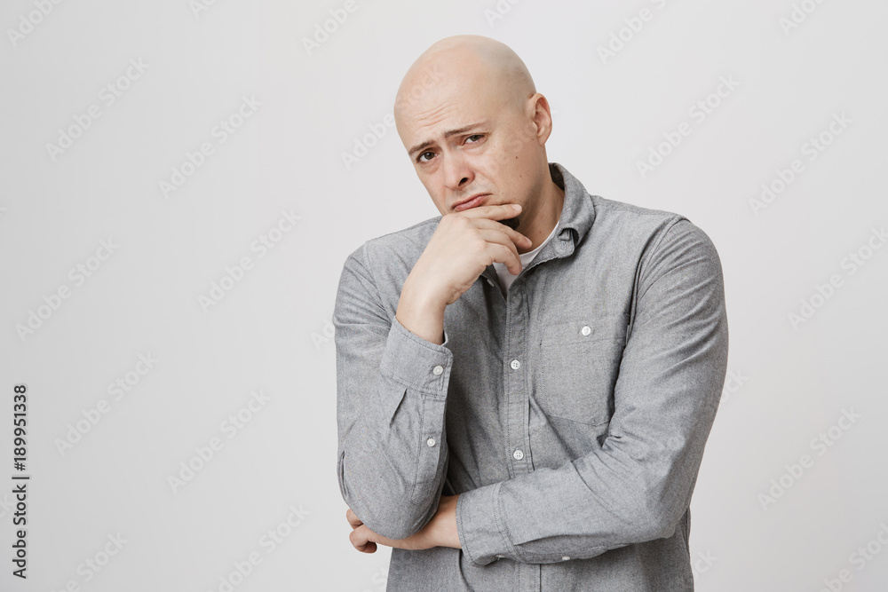 Bald businessman with beard keeps hand on chin, looks pensively at camera, frowns, isolated against gray background. Confident hairless european guy with thoughtful expression of face. Bald businessman with beard keeps hand on chin, looks pensively at camera, frowns, isolated against gray background. Confident hairless european guy with thoughtful expression of face.