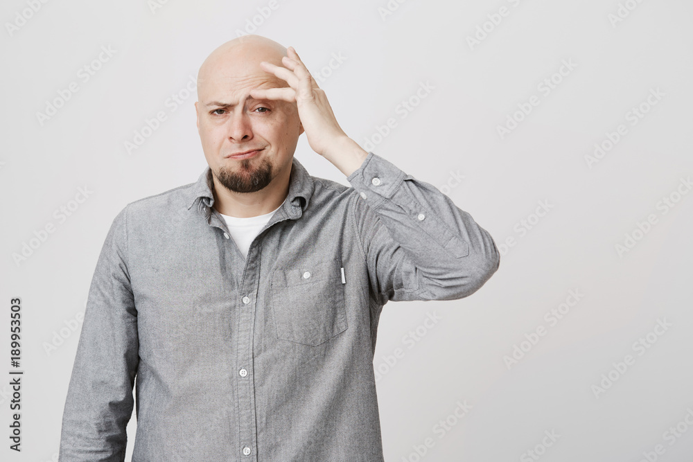 Stressed frustrated caucasian bearded bald male dressed in gray shirt touching forehead with hand, suffering from pain, being tired and overworked. Hairless man frowning face in irritation and pain Stressed frustrated caucasian bearded bald male dressed in gray shirt touching forehead with hand, suffering from pain, being tired and overworked. Hairless man frowning face in irritation and pain