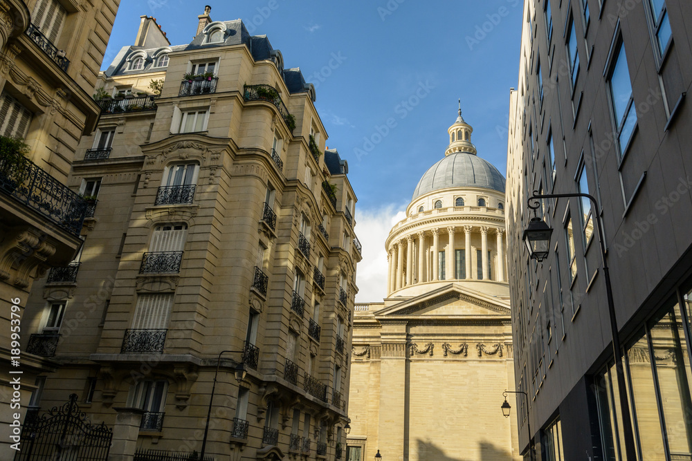 Low angle side view of the cupola of the Pantheon in Paris from a narrow adjacent street with modern and classic parisian buildings in the foreground.