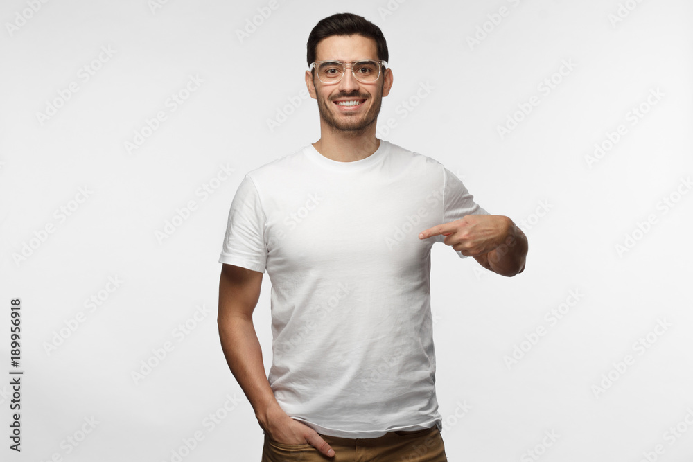 Attractive young man in transparent glasses pointing to his blank white tshirt with index finger, showing empty space for your advertising text or logo, statning isolated on grey background Attractive young man in transparent glasses pointing to his blank white tshirt with index finger, showing empty space for your advertising text or logo, statning isolated on grey background
