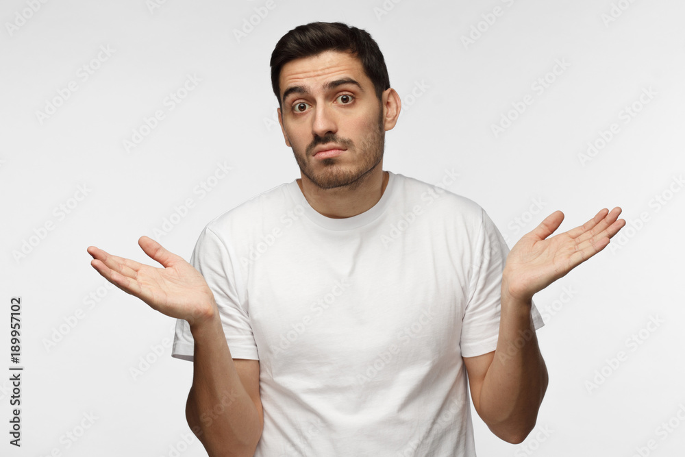 I don't know. Young man isolated on grey background being at a loss, showing helpless gesture with arm and hands, mouth curved as if he does not know what to do I don't know. Young man isolated on grey background being at a loss, showing helpless gesture with arm and hands, mouth curved as if he does not know what to do