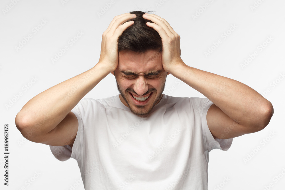 Upset unhappy young man in white t-shirt squeezing head with hands, suffering from headache. People, stress, tension and migraine concept Upset unhappy young man in white t-shirt squeezing head with hands, suffering from headache. People, stress, tension and migraine concept