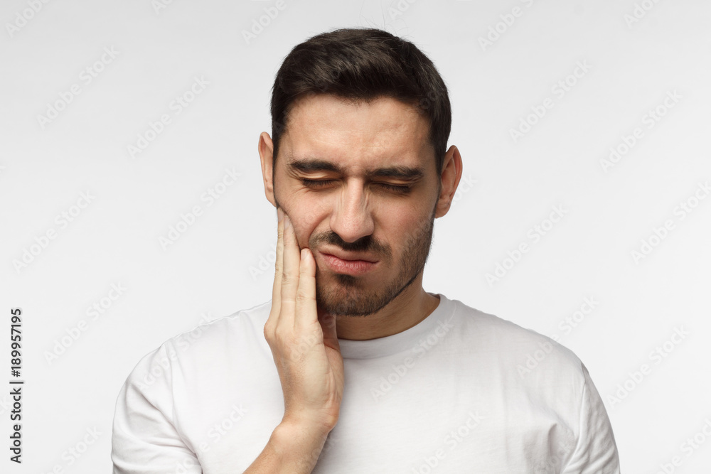 Indoor headshot of young man feeling pain, holding his cheek with hand, suffering from bad toothache, looking at camera with painful expression. Tooth ache concept. Indoor headshot of young man feeling pain, holding his cheek with hand, suffering from bad toothache, looking at camera with painful expression. Tooth ache concept.
