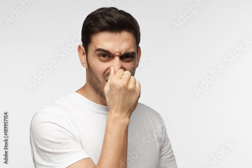 Portrait of young handsome man in casual clothes isolated on grey background holding his nose as if smelling something rotten and stinky looking aside trying to find source of odor