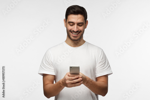 Closeup photo of man in white tshirt standing isolated on gray background looking attentively at screen of cellphone, browsing web pages and smiling nicely while chatting with friends