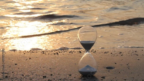 Hourglass on the beach with sea waves in bright and warm summer golden morning light showing starting time for a new day and running out in every moment
