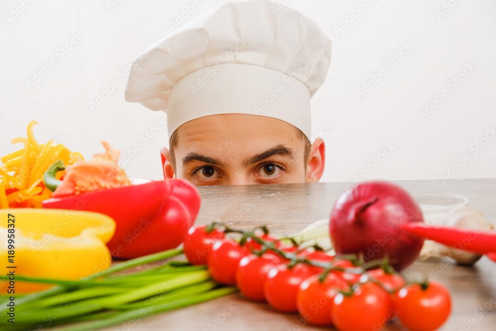 Man with cook cap on white background. Chef with vegetables on table. Cook with cheerful face in face close up. Man with cook cap on white background. Chef with vegetables on table. Cook with cheerful face in face close up.