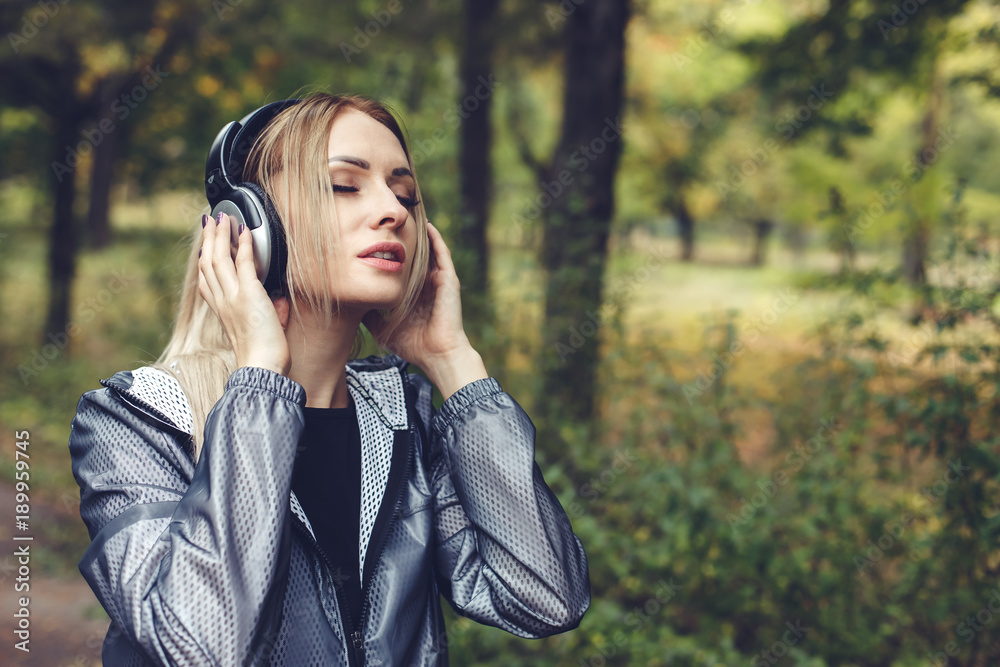 Portrait of young attractive blonde woman on a city park,  listening to music on headphones