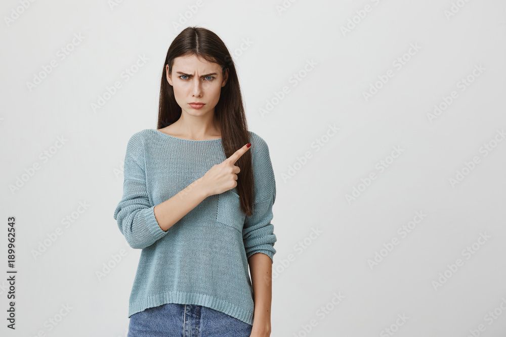 Portrait of displeased dissatisfied young woman with long dark straight hair wearing long sleeved blue sweater frowning face looking angrily at camera, pointing with forefinger at copy space. Portrait of displeased dissatisfied young woman with long dark straight hair wearing long sleeved blue sweater frowning face looking angrily at camera, pointing with forefinger at copy space.