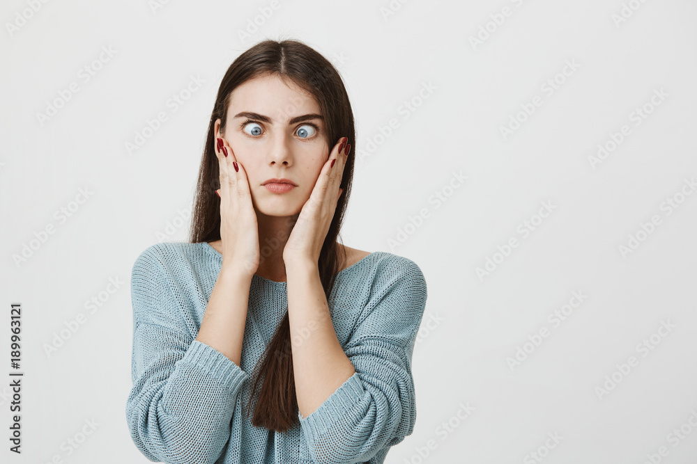 Indoor shot of pretty brunette woman making grimace with slanting blue eyes, touching her cheeks with hands, having tired and annoyed expression. Face expression and negative emotions Indoor shot of pretty brunette woman making grimace with slanting blue eyes, touching her cheeks with hands, having tired and annoyed expression. Face expression and negative emotions