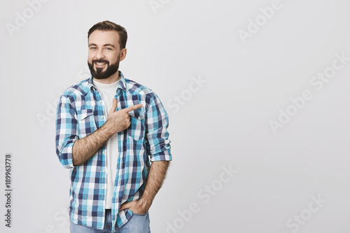 Charming bearded adult man in blue plaid shirt pointing right with index finger while another hand in pocket, over gray background. Guy works as a model for advertising new promos of store photo