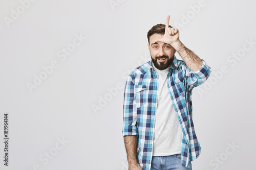 Tableau sur toile Portrait of european guy in plaid shirt holding hand on forehead showing loser gesture, smiling at camera and standing against gray background