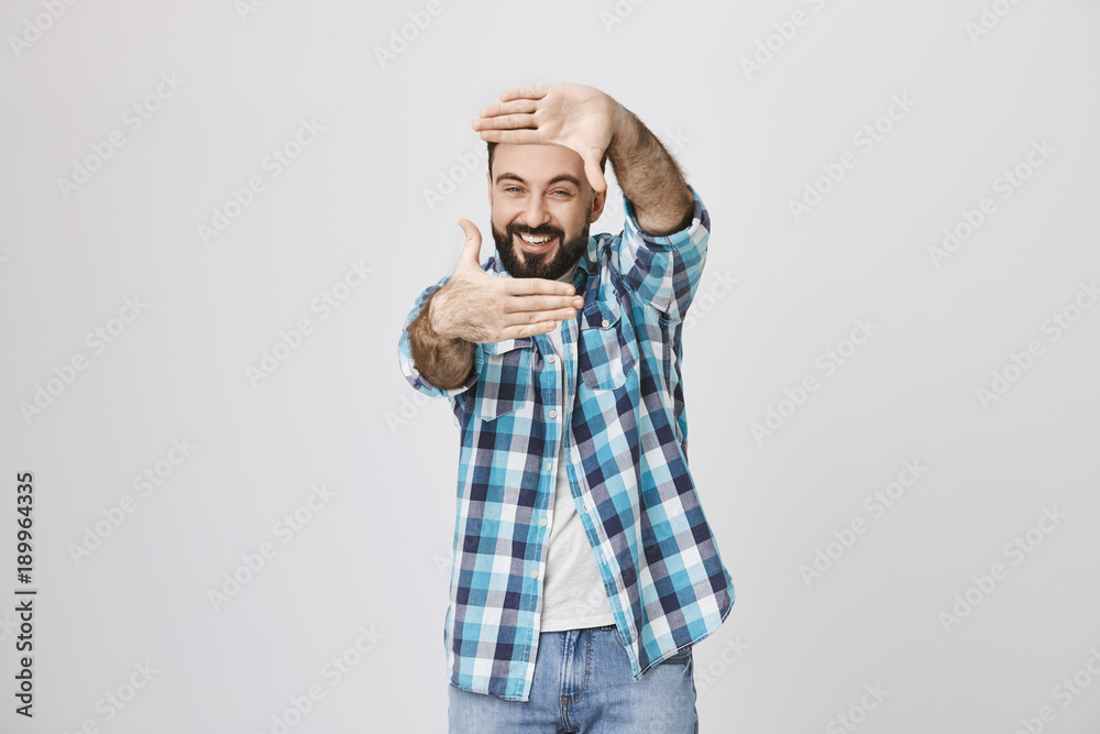 Studio shot of funny bearded european male office worker, holding hands in frame gesture while looking through it and smiling broadly, over gray background. Person acts like he is movie director Studio shot of funny bearded european male office worker, holding hands in frame gesture while looking through it and smiling broadly, over gray background. Person acts like he is movie director