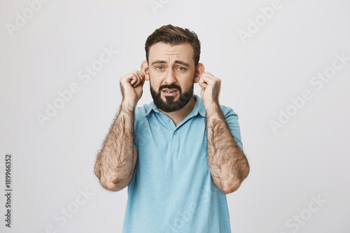 Adult bearded man with gloomy smile stretching ears, imitating sad monkey, over gray background. Guy is tired of hearing lies from his close friends. Fed up of listening demands