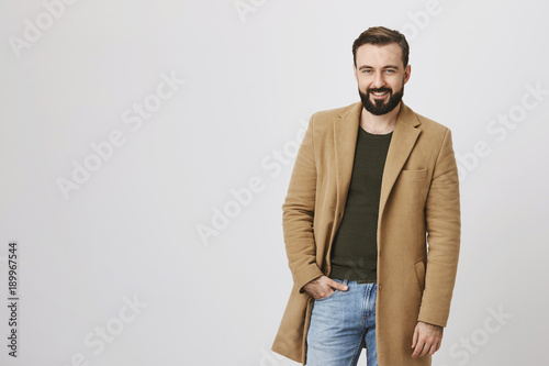 Handsome macho man with beard and moustache, wearing light-brown coat over green pullover, holding hand in jeans, expressing confidence while standing against gray background. photo