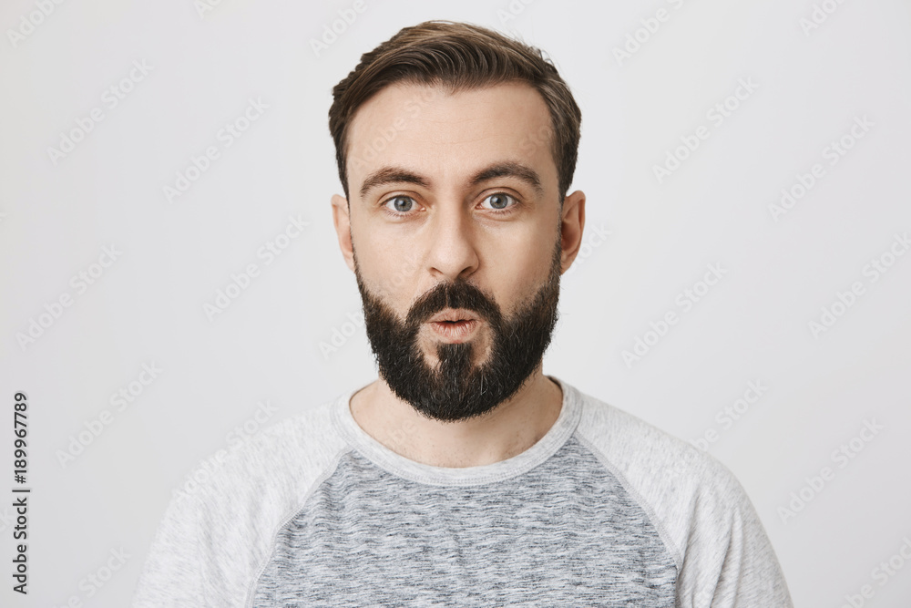 Indoor portrait of amazed bearded guy saying wow while looking at camera and standing against gray background. Man amazed by new gadget he sees in tv promotion thinking about ordering it Indoor portrait of amazed bearded guy saying wow while looking at camera and standing against gray background. Man amazed by new gadget he sees in tv promotion thinking about ordering it