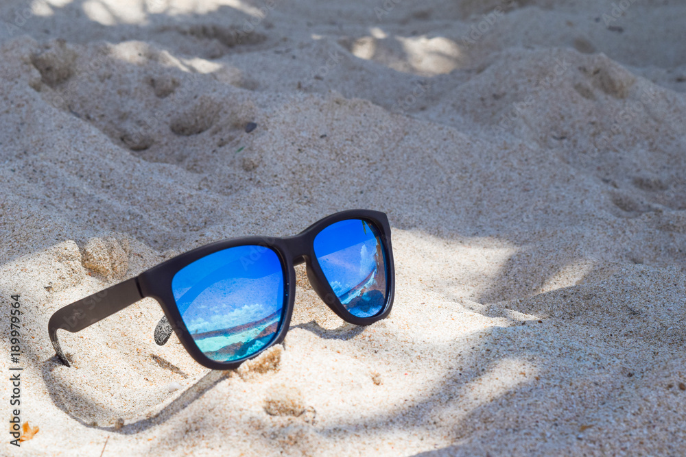 Sonnenbrille im weißen Sand, in den Gläsern spiegelt sich der blaue Himmel und das türkis-farbene Meer