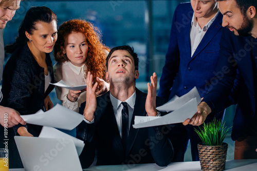 stressed male worker raises his hands and says please God while his colleagues ask him to do the job