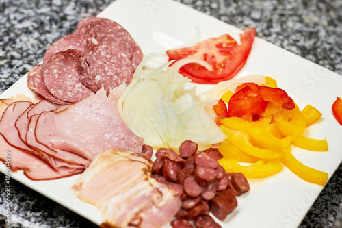 Sliced cold meats and vegetables on white plate. Pizza ingredients assortment, close up. Food assortment.