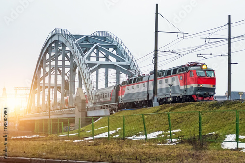 High speed red passenger train rushing through the bridge industrial railway in the evening at sunset.