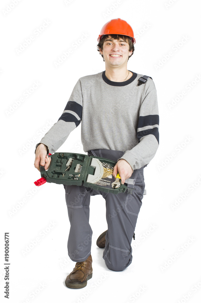A young man in working grey clothes and orange hard helmet sitting with toolkit on isolated white background. A young man in working grey clothes and orange hard helmet sitting with toolkit on isolated white background.