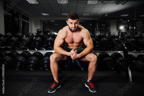 bearded young bodybuilder sits on a stack with dumbbells. Rest between exercises.