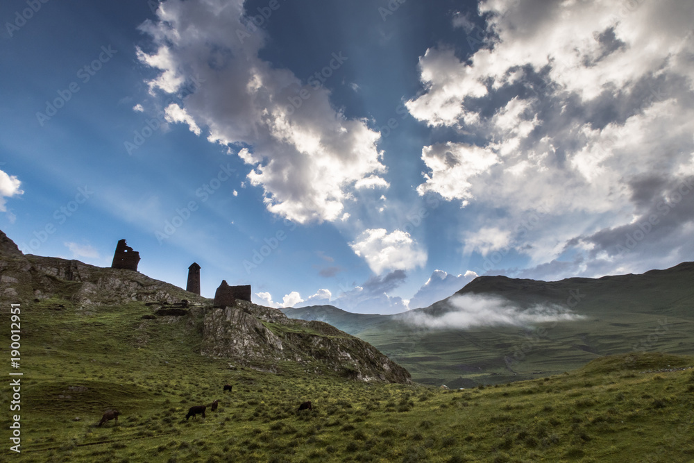Summer morning at Caucasus, Tusheti region , Georgia. Hills covered with green grass. On the high rocks located ruins of medival tushetian defensive tower. Sunrays  highlighting clouds in the blue sky