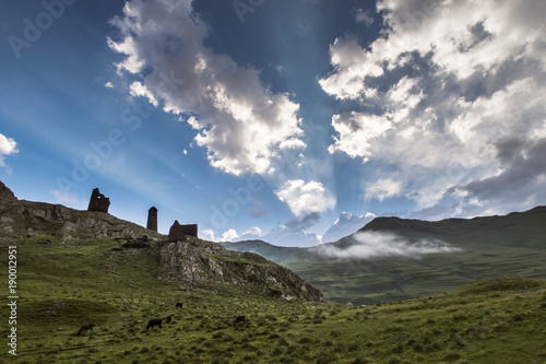 Summer morning at Caucasus  Tusheti region   Georgia. Hills covered with green grass. On the high rocks located ruins of medival tushetian defensive tower. Sunrays  highlighting clouds in the blue sky