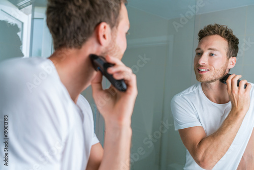 Shaving beard with electric shaver in bathroom. Man looking in mirror, male beauty care routine lifestyle.
