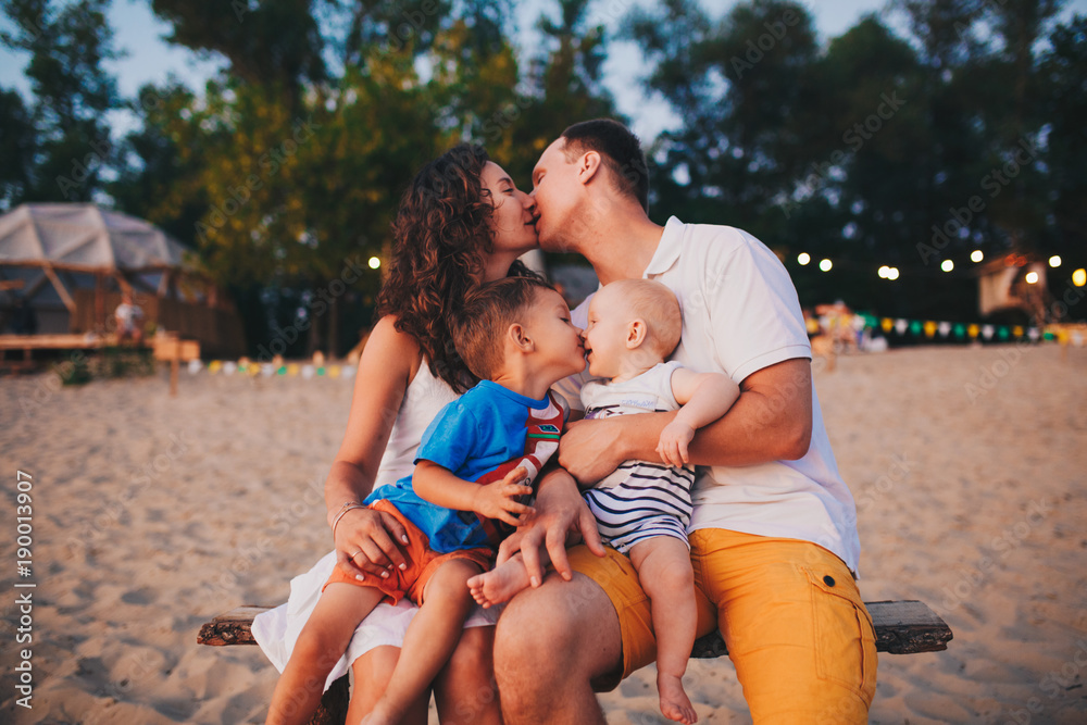 Fototapeta premium The concept of a family vacation. Young family and two sons sitting on a bench in the evening on a sandy beach. Mom and Dad kiss, the older brother kisses the younger on the lips