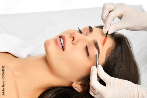 Beautician doing permanent eyebrows makeup. Closeup composition of beautiful woman face with thick brows In beauty salon photo