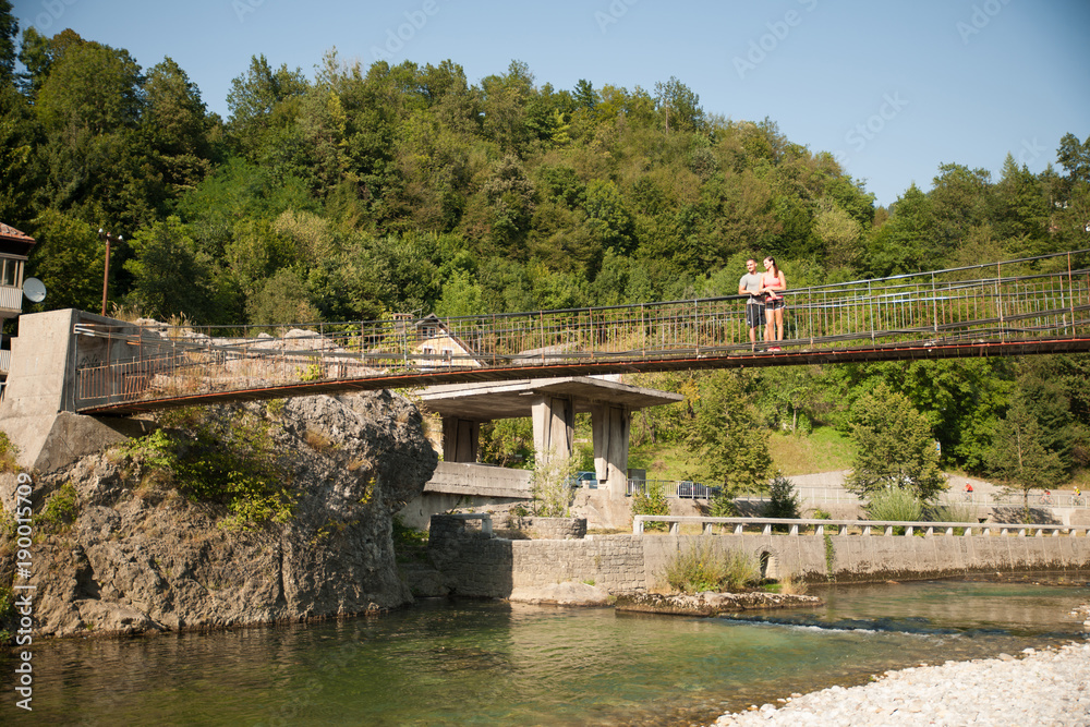 Active young couple on a bridge over alpine river on hot summer morning