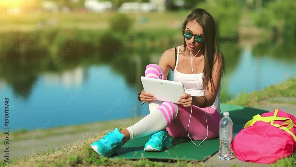 Girl sits near pond and talks via tablet computer, sportswoman talks on tablet pc. Woman in sportswear with tablet pc communicates through skype or video chat, beautiful morning sun at background