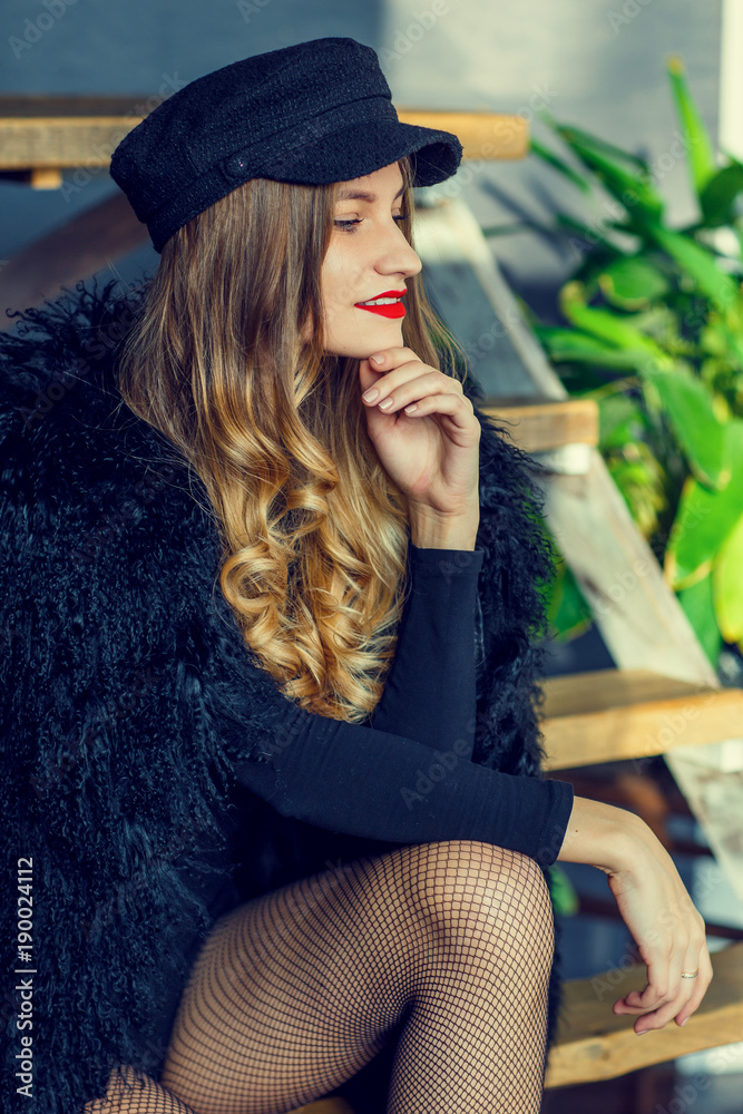 In-Studio lifestyle portrait of a stunning fashionable girl in the cap. Smiling, sitting on the wooden stairs. Trendsetter. In a stylish black coat from Lama and fishnet tights. In-Studio lifestyle portrait of a stunning fashionable girl in the cap. Smiling, sitting on the wooden stairs. Trendsetter. In a stylish black coat from Lama and fishnet tights.
