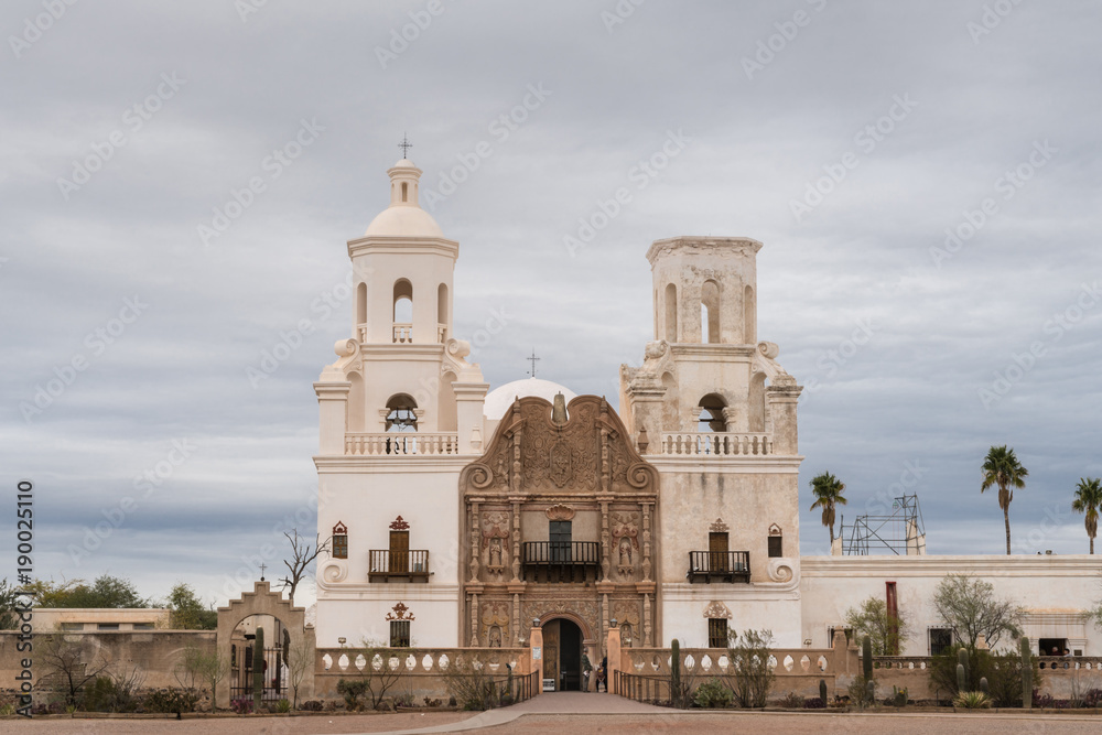 Tucson, Arizona, USA - January 9, 2018: White and brown front facade of Historic San Xavier Del Bac Mission under heavy gray, white cloud deck. Some greenish desert plants in front.