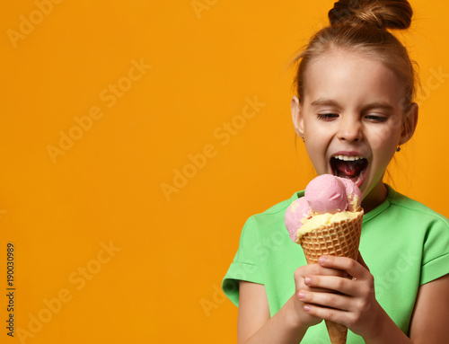 Fototapet Pretty baby girl kid eating licking banana and strawberry ice cream in waffles c