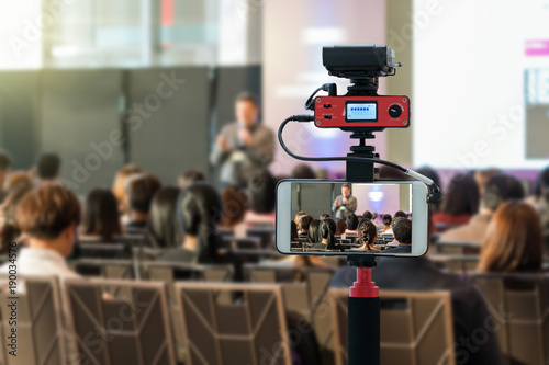 Closeup smart mobile phone taking Live over Speakers on the stage with Rear view of Audience in the conference hall or seminar meeting, technology live streaming and broadcast concept photo