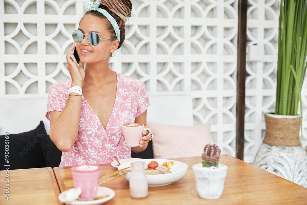 Stylish female wears shades, looks aside, holds cup of delicious espresso with foam, has pleasant conversation via modern cell phone. Happy young woman calls during morning breakfast at cafeteria Stylish female wears shades, looks aside, holds cup of delicious espresso with foam, has pleasant conversation via modern cell phone. Happy young woman calls during morning breakfast at cafeteria