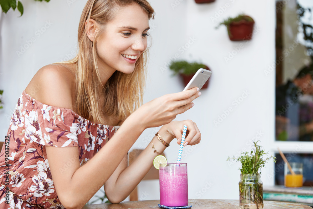 Happy beautiful young woman in stylish blouse, sends messages to friends on cell phone, congrtulates them with holiday, drinks smoothie drink with straw, spends spare time at cozy resaturant Happy beautiful young woman in stylish blouse, sends messages to friends on cell phone, congrtulates them with holiday, drinks smoothie drink with straw, spends spare time at cozy resaturant