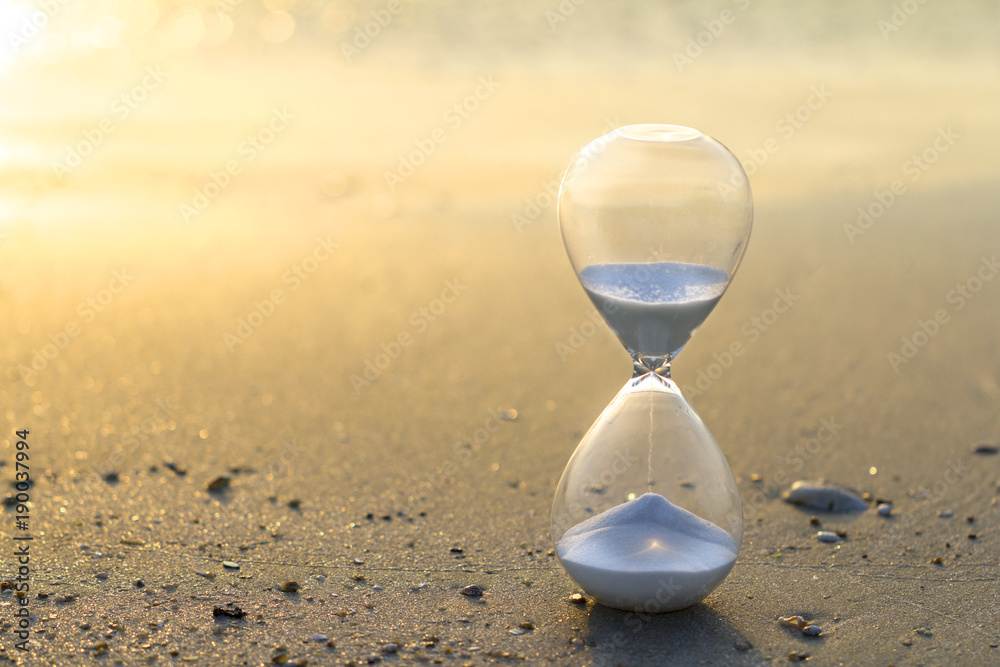 Hourglass close up in a warm golden morning sunlight on a sandy beach starting time for a new day or running of time with side copy space. Hourglass close up in a warm golden morning sunlight on a sandy beach starting time for a new day or running of time with side copy space.