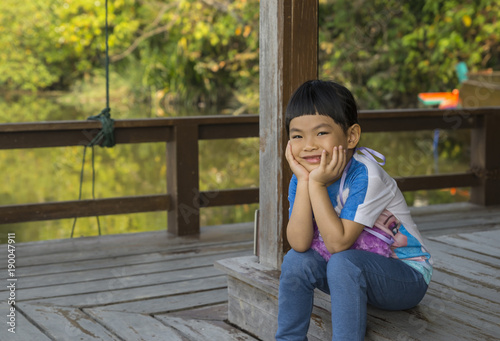  Cute little Asian girl with big smile sitting on the wood floor of the riverside pavilion. Hands on chin and look at camera. Happy time in holiday with family. Wooden riverside pavilion in Thailand.