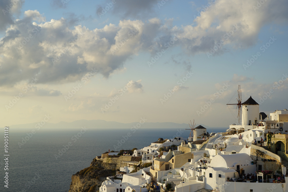 Fototapeta premium Beautiful evening light scene of Oia white building townscape and windmill along island mountain, vast Aegean sea, abstract cloud and blue sky background