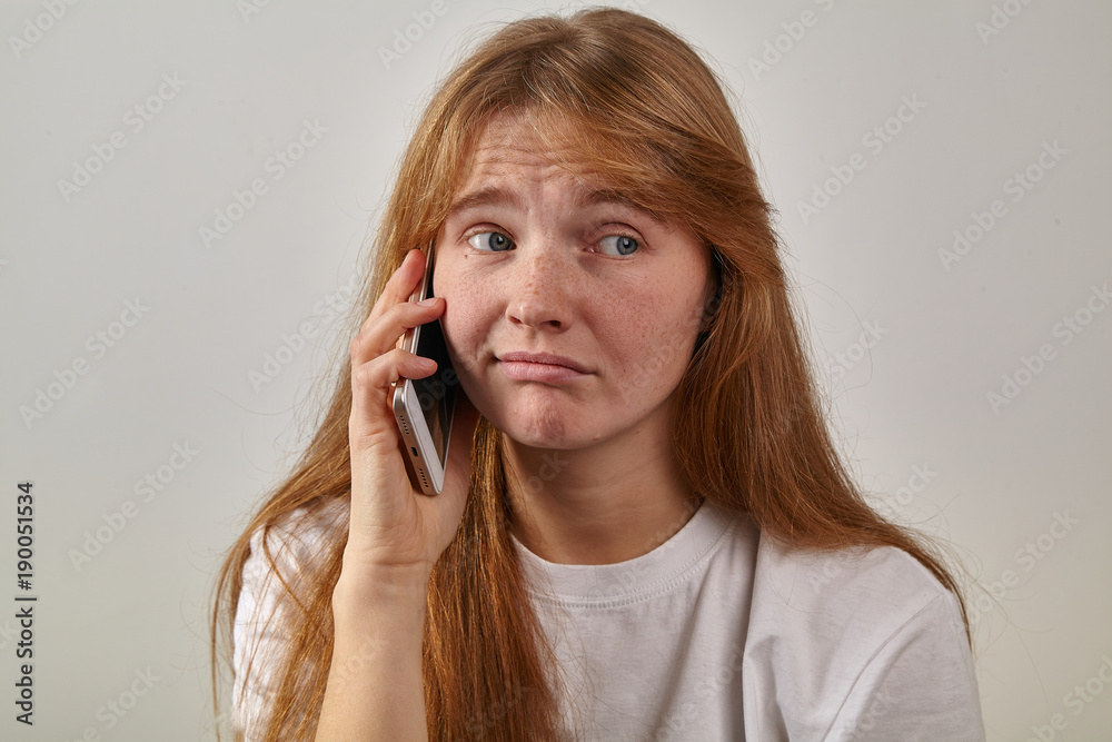Headshot of young red-headed girl with freckles holding her phone next to her ear and talking with sad expression on her face Headshot of young red-headed girl with freckles holding her phone next to her ear and talking with sad expression on her face