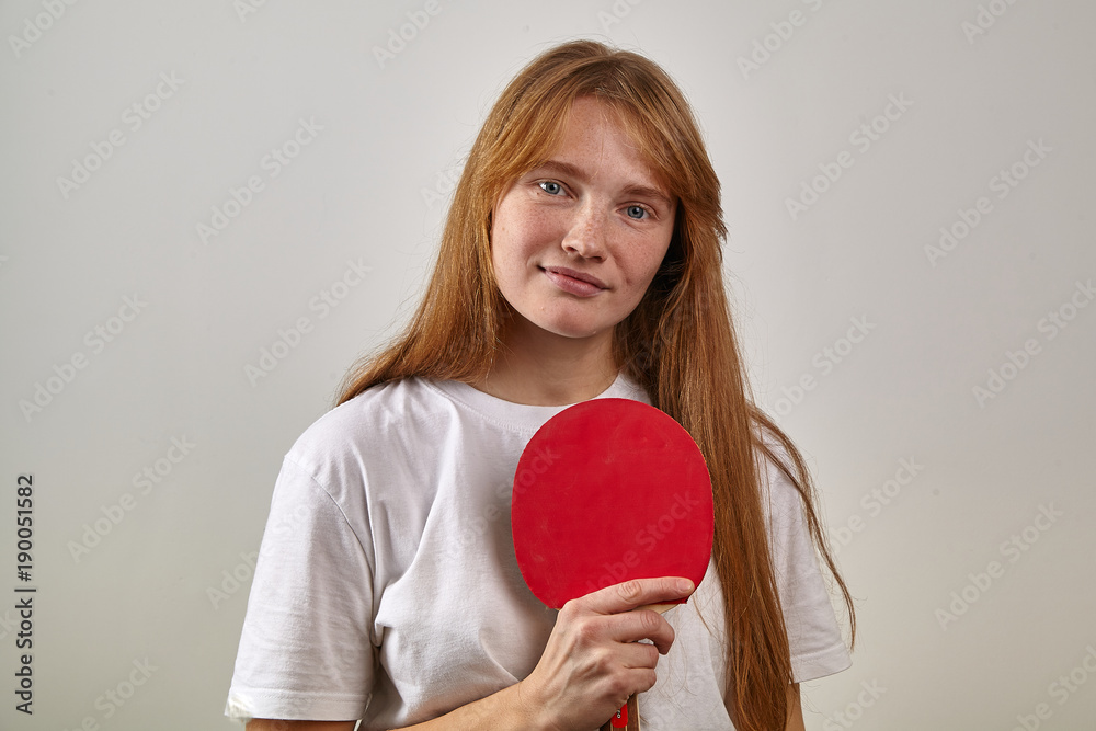 Studio indoor portrait of young girl with red hair and freckles dressed in white t-shirt holding table tennis racket and looking at the camera Studio indoor portrait of young girl with red hair and freckles dressed in white t-shirt holding table tennis racket and looking at the camera